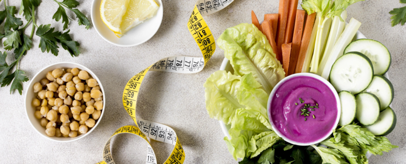 Widok z góry na kompozycję zdrowego posiłku i akcesoriów dietetycznych na jasnym, teksturowanym tle. W centrum znajduje się biała, prostokątna taca wypełniona różnorodnymi pokrojonymi warzywami: chrupiącymi liśćmi sałaty, pałeczkami marchewki i selera naciowego, oraz plasterkami ogórka. Na środku tacy umieszczony jest mały, biały kubeczek z intensywnie fioletowym dipem (prawdopodobnie na bazie buraków), posypanym świeżym szczypiorkiem. Obok, w osobnej białej miseczce, leży ugotowana ciecierzyca. Na białym spodku widoczne są świeże plasterki cytryny. Całość kompozycji otacza swobodnie zawinięta żółta miarka krawiecka, a uzupełniają ją zielone gałązki natki pietruszki