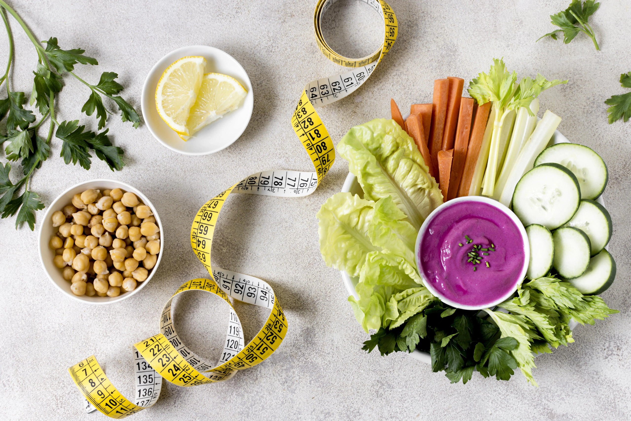 Widok z góry na kompozycję zdrowego posiłku i akcesoriów dietetycznych na jasnym, teksturowanym tle. W centrum znajduje się biała, prostokątna taca wypełniona różnorodnymi pokrojonymi warzywami: chrupiącymi liśćmi sałaty, pałeczkami marchewki i selera naciowego, oraz plasterkami ogórka. Na środku tacy umieszczony jest mały, biały kubeczek z intensywnie fioletowym dipem (prawdopodobnie na bazie buraków), posypanym świeżym szczypiorkiem. Obok, w osobnej białej miseczce, leży ugotowana ciecierzyca. Na białym spodku widoczne są świeże plasterki cytryny. Całość kompozycji otacza swobodnie zawinięta żółta miarka krawiecka, a uzupełniają ją zielone gałązki natki pietruszki