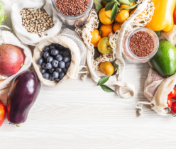 Zdjęcie z góry (flat lay) przedstawiające bogactwo świeżych produktów spożywczych i suchych towarów, ułożonych na jasnym drewnianym blacie. Produkty są częściowo zapakowane w ekologiczne, siatkowe i płócienne torby wielokrotnego użytku oraz szklane słoiki, co sugeruje podejście zero-waste. Widoczne są m.in.: worki z ciemnymi borówkami, mango, jasnymi nasionami (prawdopodobnie grochem lub fasolą), a także szklane słoiki z brązowym ryżem i czerwoną soczewicą. Pośród nich leżą świeże owoce, takie jak mandarynki (niektóre z liśćmi), małe kumkwaty oraz fragmenty bananów. Wśród warzyw wyróżniają się: fioletowy bakłażan, czerwona i żółta papryka, zielona sałata (lub kapusta), dojrzałe awokado oraz kiść czerwonych pomidorków koktajlowych. Całość kompozycji tworzy obraz zdrowego i zrównoważonego stylu życia