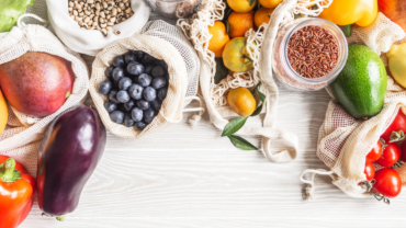 Zdjęcie z góry (flat lay) przedstawiające bogactwo świeżych produktów spożywczych i suchych towarów, ułożonych na jasnym drewnianym blacie. Produkty są częściowo zapakowane w ekologiczne, siatkowe i płócienne torby wielokrotnego użytku oraz szklane słoiki, co sugeruje podejście zero-waste. Widoczne są m.in.: worki z ciemnymi borówkami, mango, jasnymi nasionami (prawdopodobnie grochem lub fasolą), a także szklane słoiki z brązowym ryżem i czerwoną soczewicą. Pośród nich leżą świeże owoce, takie jak mandarynki (niektóre z liśćmi), małe kumkwaty oraz fragmenty bananów. Wśród warzyw wyróżniają się: fioletowy bakłażan, czerwona i żółta papryka, zielona sałata (lub kapusta), dojrzałe awokado oraz kiść czerwonych pomidorków koktajlowych. Całość kompozycji tworzy obraz zdrowego i zrównoważonego stylu życia