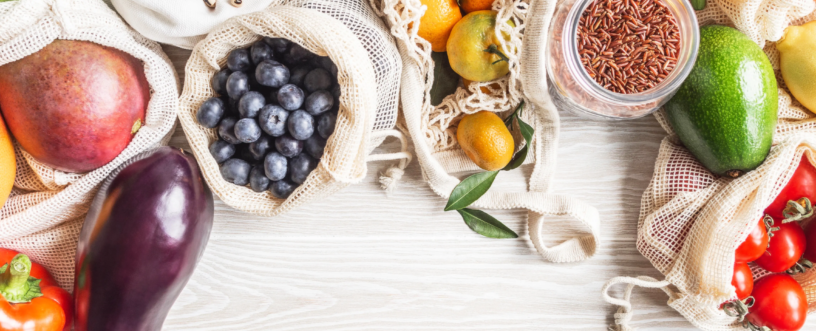 Zdjęcie z góry (flat lay) przedstawiające bogactwo świeżych produktów spożywczych i suchych towarów, ułożonych na jasnym drewnianym blacie. Produkty są częściowo zapakowane w ekologiczne, siatkowe i płócienne torby wielokrotnego użytku oraz szklane słoiki, co sugeruje podejście zero-waste. Widoczne są m.in.: worki z ciemnymi borówkami, mango, jasnymi nasionami (prawdopodobnie grochem lub fasolą), a także szklane słoiki z brązowym ryżem i czerwoną soczewicą. Pośród nich leżą świeże owoce, takie jak mandarynki (niektóre z liśćmi), małe kumkwaty oraz fragmenty bananów. Wśród warzyw wyróżniają się: fioletowy bakłażan, czerwona i żółta papryka, zielona sałata (lub kapusta), dojrzałe awokado oraz kiść czerwonych pomidorków koktajlowych. Całość kompozycji tworzy obraz zdrowego i zrównoważonego stylu życia