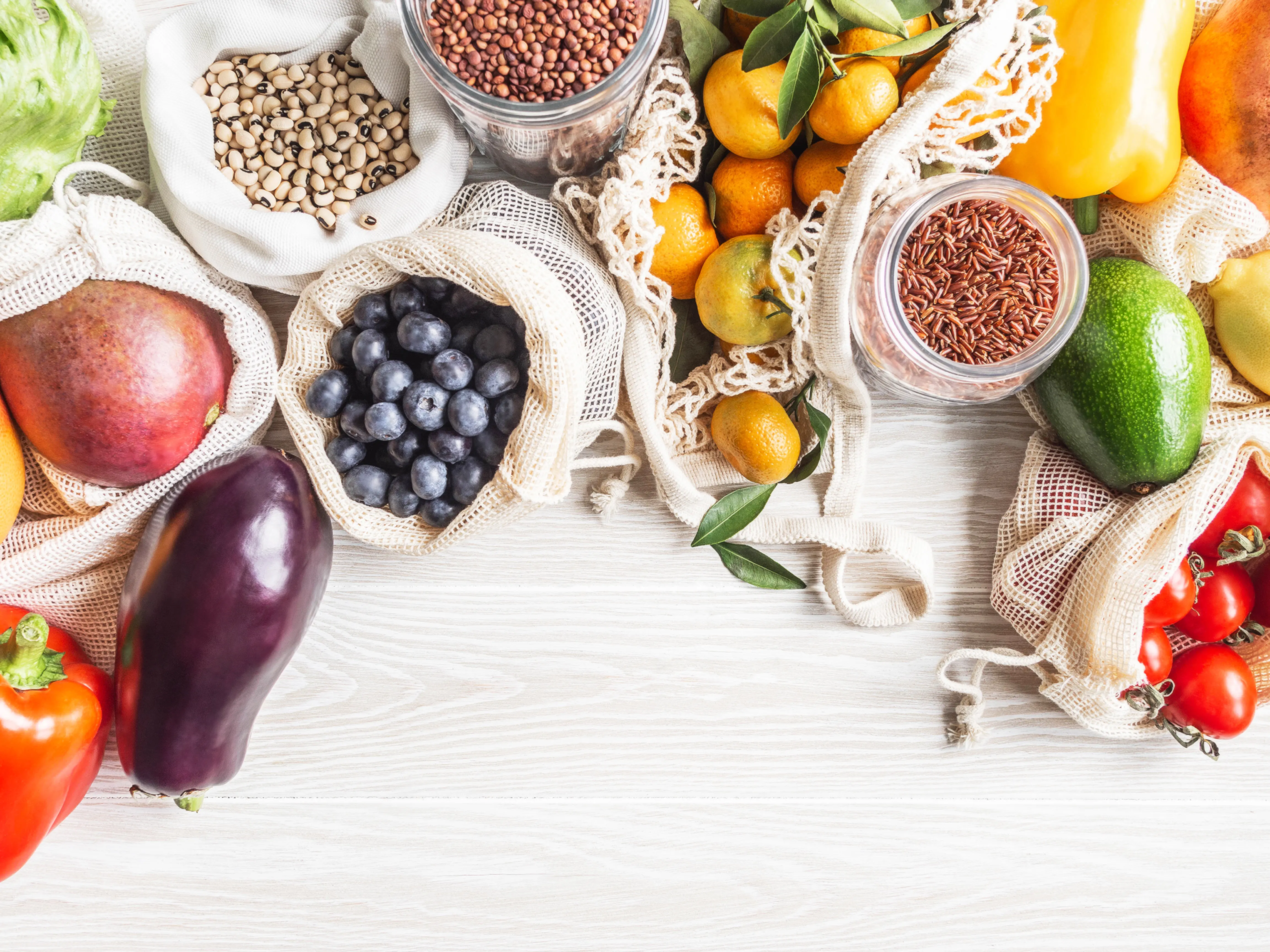 Zdjęcie z góry (flat lay) przedstawiające bogactwo świeżych produktów spożywczych i suchych towarów, ułożonych na jasnym drewnianym blacie. Produkty są częściowo zapakowane w ekologiczne, siatkowe i płócienne torby wielokrotnego użytku oraz szklane słoiki, co sugeruje podejście zero-waste. Widoczne są m.in.: worki z ciemnymi borówkami, mango, jasnymi nasionami (prawdopodobnie grochem lub fasolą), a także szklane słoiki z brązowym ryżem i czerwoną soczewicą. Pośród nich leżą świeże owoce, takie jak mandarynki (niektóre z liśćmi), małe kumkwaty oraz fragmenty bananów. Wśród warzyw wyróżniają się: fioletowy bakłażan, czerwona i żółta papryka, zielona sałata (lub kapusta), dojrzałe awokado oraz kiść czerwonych pomidorków koktajlowych. Całość kompozycji tworzy obraz zdrowego i zrównoważonego stylu życia