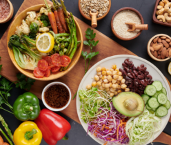 Zdjęcie z góry (flat lay) prezentujące obfite, zdrowe posiłki wegańskie lub wegetariańskie, ułożone na drewnianej desce i ciemnym tle. Po lewej stronie, w drewnianej misce, znajduje się kompozycja pieczonych warzyw, takich jak kalafior, brokuły, marchewki, szparagi i pak choi, uzupełniona świeżymi pomidorkami koktajlowymi i plastrem cytryny. W małej miseczce obok jest ciemny sos z sezamem. Po prawej stronie, na białym talerzu, widnieje sałatka z ciecierzycą, czerwoną fasolą, plastrami ogórka, połówką awokado, kiełkami oraz szatkowaną czerwoną i białą kapustą z marchewką. Wokół głównych dań rozmieszczone są mniejsze, drewniane miseczki z dodatkami: różową solą himalajską, różnokolorowym pieprzem, pestkami słonecznika, sezamem, orzechami nerkowca i migdałami. Na ciemnym blacie widać także świeże, całe papryki (zieloną, żółtą, czerwoną), szparagi, małe marchewki, natkę pietruszki i przekrojone cytryny