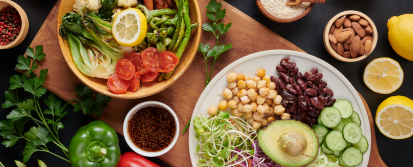 Zdjęcie z góry (flat lay) prezentujące obfite, zdrowe posiłki wegańskie lub wegetariańskie, ułożone na drewnianej desce i ciemnym tle. Po lewej stronie, w drewnianej misce, znajduje się kompozycja pieczonych warzyw, takich jak kalafior, brokuły, marchewki, szparagi i pak choi, uzupełniona świeżymi pomidorkami koktajlowymi i plastrem cytryny. W małej miseczce obok jest ciemny sos z sezamem. Po prawej stronie, na białym talerzu, widnieje sałatka z ciecierzycą, czerwoną fasolą, plastrami ogórka, połówką awokado, kiełkami oraz szatkowaną czerwoną i białą kapustą z marchewką. Wokół głównych dań rozmieszczone są mniejsze, drewniane miseczki z dodatkami: różową solą himalajską, różnokolorowym pieprzem, pestkami słonecznika, sezamem, orzechami nerkowca i migdałami. Na ciemnym blacie widać także świeże, całe papryki (zieloną, żółtą, czerwoną), szparagi, małe marchewki, natkę pietruszki i przekrojone cytryny