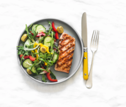 Zdjęcie z góry przedstawiające zdrowy i kolorowy posiłek składający się z grillowanego łososia i świeżej sałatki, ułożonych na okrągłym, szarym talerzu. Łosoś jest złocisty, z wyraźnymi śladami grillowania. Sałatka zawiera zieloną rukolę, świeże plasterki ogórka, paski czerwonej papryki, grillowane kawałki żółtej papryki, zielone oliwki oraz cienkie krążki białej cebuli. Obok talerza leżą srebrne sztućce: widelec i nóż z rączką w intensywnym, musztardowym kolorze. Całość ułożona jest na jasnym, lekko pogniecionym, teksturowanym tle przypominającym obrus