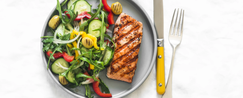 Zdjęcie z góry przedstawiające zdrowy i kolorowy posiłek składający się z grillowanego łososia i świeżej sałatki, ułożonych na okrągłym, szarym talerzu. Łosoś jest złocisty, z wyraźnymi śladami grillowania. Sałatka zawiera zieloną rukolę, świeże plasterki ogórka, paski czerwonej papryki, grillowane kawałki żółtej papryki, zielone oliwki oraz cienkie krążki białej cebuli. Obok talerza leżą srebrne sztućce: widelec i nóż z rączką w intensywnym, musztardowym kolorze. Całość ułożona jest na jasnym, lekko pogniecionym, teksturowanym tle przypominającym obrus