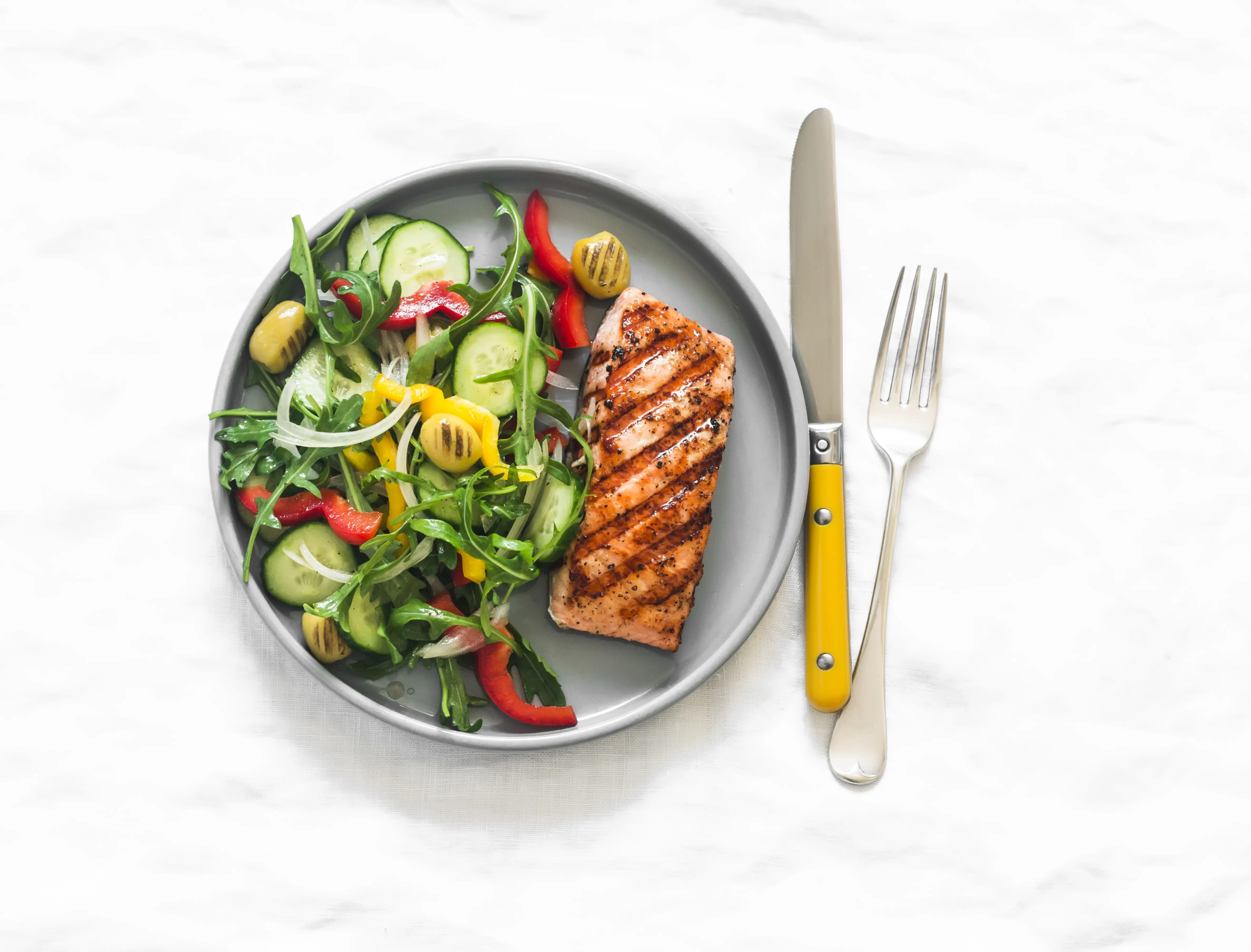 Zdjęcie z góry przedstawiające zdrowy i kolorowy posiłek składający się z grillowanego łososia i świeżej sałatki, ułożonych na okrągłym, szarym talerzu. Łosoś jest złocisty, z wyraźnymi śladami grillowania. Sałatka zawiera zieloną rukolę, świeże plasterki ogórka, paski czerwonej papryki, grillowane kawałki żółtej papryki, zielone oliwki oraz cienkie krążki białej cebuli. Obok talerza leżą srebrne sztućce: widelec i nóż z rączką w intensywnym, musztardowym kolorze. Całość ułożona jest na jasnym, lekko pogniecionym, teksturowanym tle przypominającym obrus